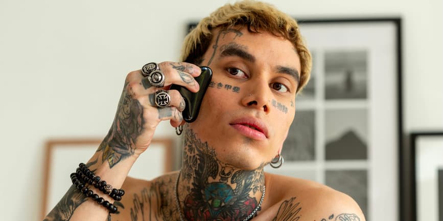 Gua Sha stone being used on face Portrait of a heavily tattooed man with light brown skin and bleached blond hair using a gua sha stone on their face.