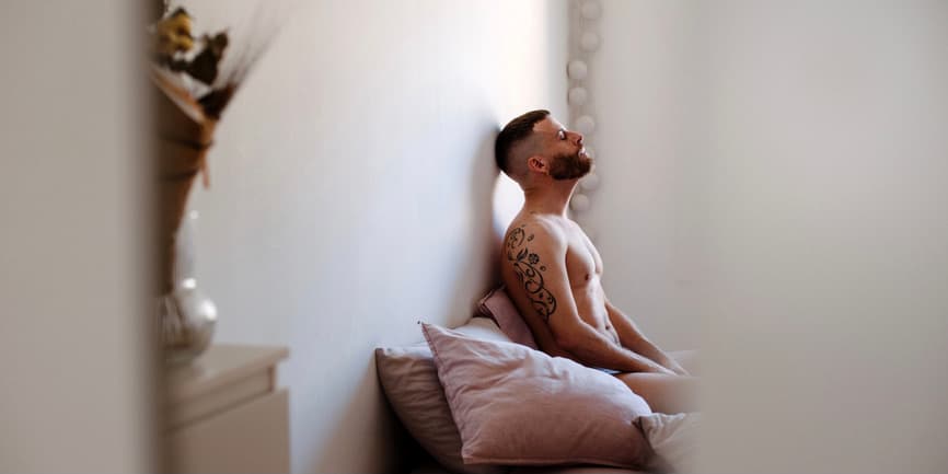 Pensive man alone in his bedroom, sitting in bed A white man with short hair and a beard wearing no shirt sits in bed with his head up against the wall. He has tattoos on his arms and is viewed through the doorframe.