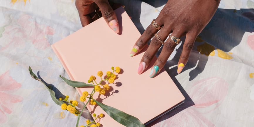 Sunlight and colorful fingernails PortraitBlack female hands with rings and colorful nails hold a pastel pink notebook on top of a floral tablecloth, with yellow flowers nearby.of stylish white senior woman wearing black and white clothing and sunglasses, laughing while she holds her pet dogs while seated on a bench at home.