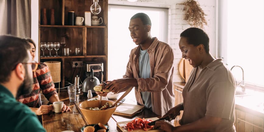 Dos parejas jóvenes sonríen, conversan y preparan la cena juntas en una cocina soleada llena de detalles en madera y piedra.