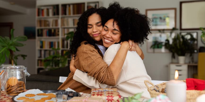Two friends hugging while making holiday cookies Close-up of two young happy Black women embracing while preparing xmas cookie boxes at home.