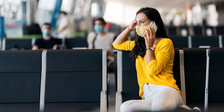 Foto de una mujer blanca de mediana edad con un suéter amarillo y una máscara facial, usando su teléfono mientras espera su vuelo en un aeropuerto.
