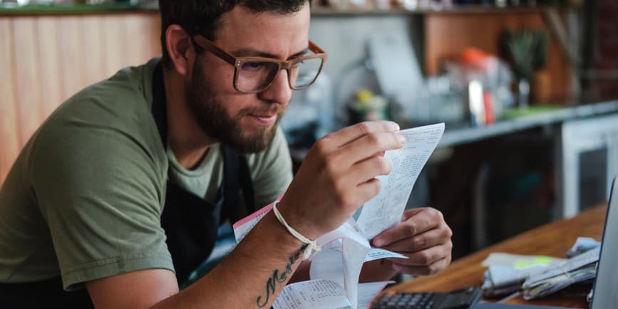 Business manager working on bills A young Latino man wearing glasses and a green t-shirt works at the counter of his business, looking intently at a receipt while working on finances.