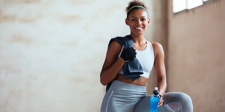 Woman at the gym A young Black woman with brown hair tied back wearing gym clothes smiles into the camera.
