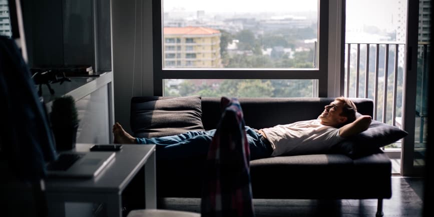 Man on couch A young white man stretched out on a couch in a modern apartment dusted in dim afternoon light, looking up at a screen.