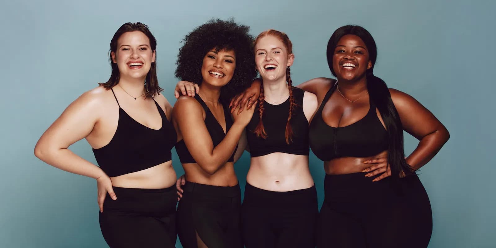 Four young women, two Black and two white with varying hair styles and body types, stand against a dark blue wall wearing black gym clothes, smiling into the camera with their arms around each other.