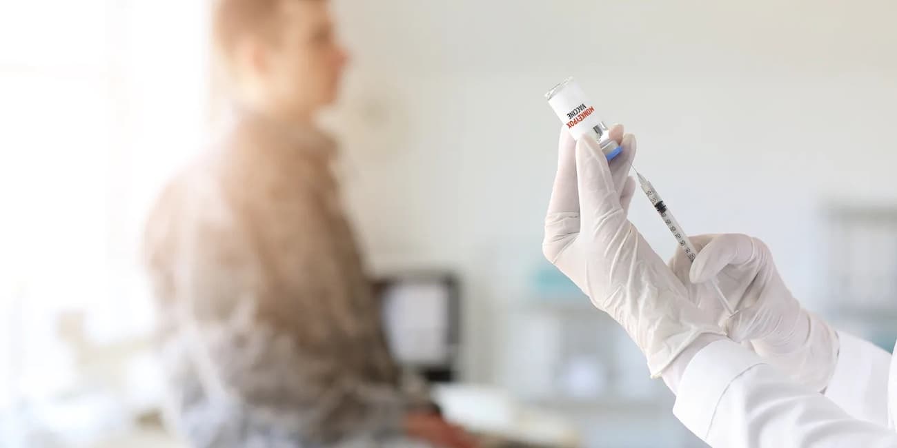 A closeup photo of a doctor with gloved hands injecting a syringe with the mpox vaccine and a blurred patient on an exam table in the background.