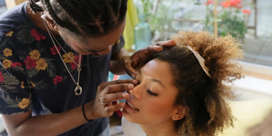 Boyfriend putting on girlfriend's lashes A handsome young Black man wearing a gray shirt topped with colorful flowers applies eyelashes to his attractive Black girlfriend, who sits with her eyes closed wearing a white t-shirt with a comb pinning back her brown Afro.