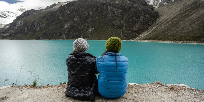 Women looking at water in winter Two women seen from behind sit on the edge of a large blue lake surrounded by gray slate mountains, wearing winter coats and hats, looking out at the water.