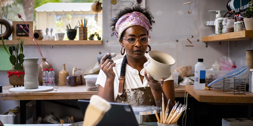 Woman painting pottery A stylish Black woman wearing a brown leather apron, glasses and violet bandana sits in a ceramics studio surrounded by instruments, concentrating on a pot in her hand.