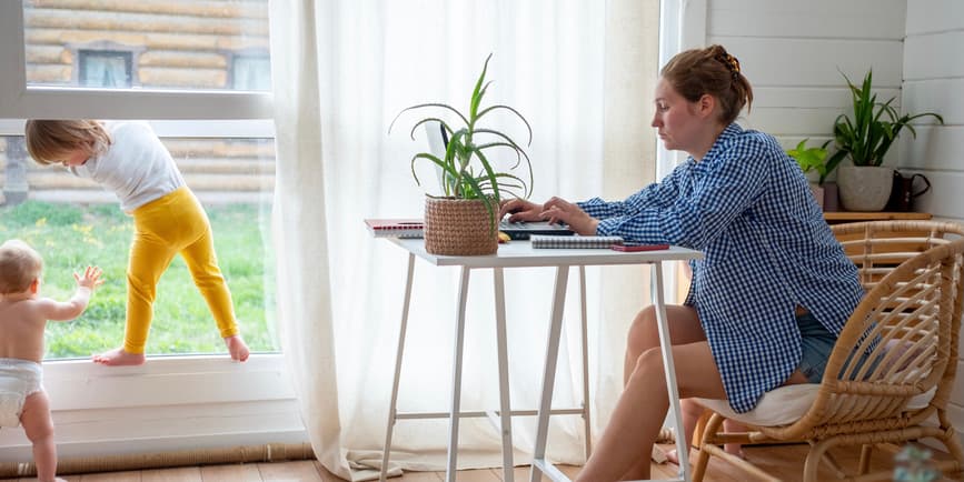 Tired mom working from home. A young white mother wearing a long sleeved shirt and no pants sits slumped in front of a computer at a small table while her toddler and baby play by the windows of her airy, modern home.