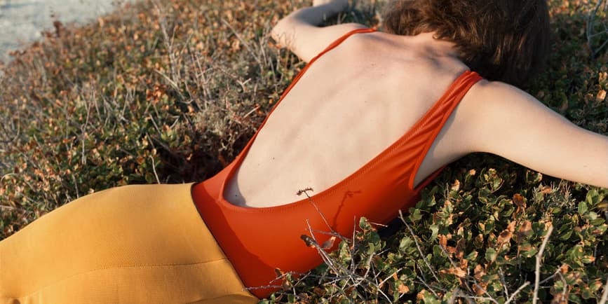 Una mujer blanca de pelo corto y rojo vestida con un body rojo oscuro sin tirantes y una falda naranja yace boca abajo en un arbusto.