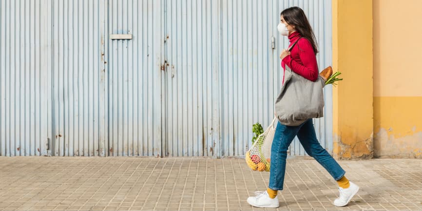 Una mujer joven y delgada de pelo largo y castaño que lleva unos vaqueros, un suéter y una máscara facial camina por una calle de la ciudad al volver de compras, con fruta fresca, verduras y pan en bolsas reutilizables.