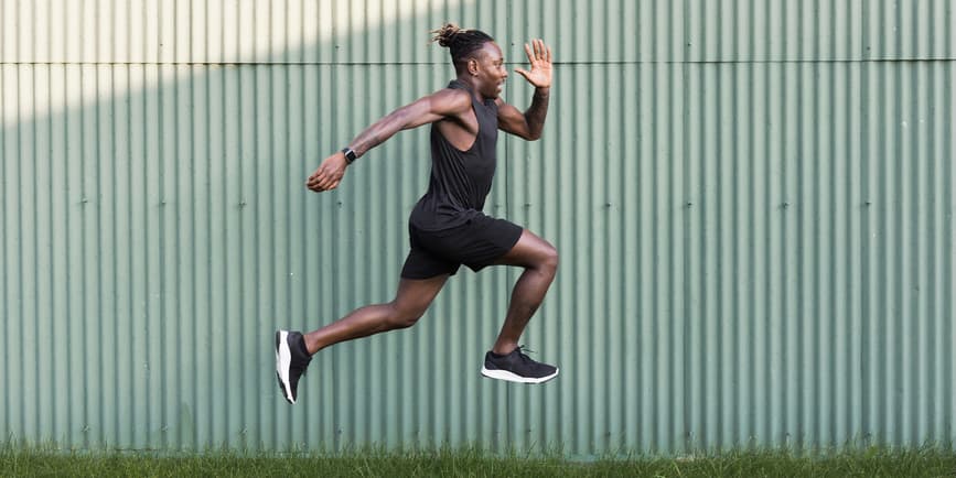 Man jumping outside Young Black man wearing workout clothes doing physical exercises by taking big jumps outdoors against a stamped metal wall with tufts of grass on the ground.