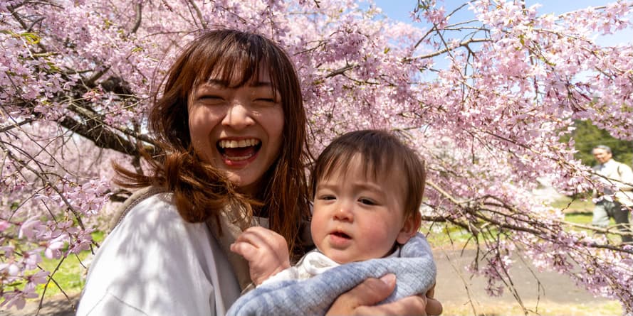 Una mujer joven de cabello castaño que lleva un jersey sonríe mientras sostiene a su hijo pequeño frente a un cerezo japonés florecido.