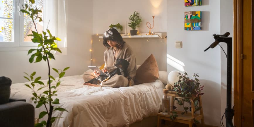 Woman on bed with dog A dreamy indoor photo of a young Asian woman wearing pajamas, sitting on her made bed in a bedroom with plants and artwork, looking down into a book as a dachshund sits on her lap and looks up at her.