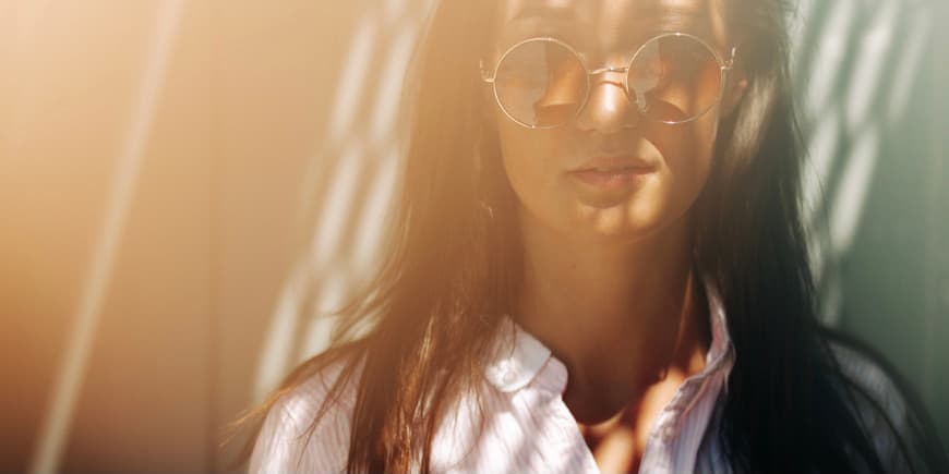 Woman in sun and shadow Portrait of a young woman with tanned skin and long dark hair wearing round sunglasses and a collared shirt standing in natural sunlight with shadows on her face.