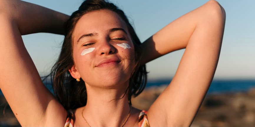 Woman in sun wearing sunblock A young white woman with short brown hair and sunscreen visible on her cheeks stands smiling in the sunset, her arms raised up and locked behind her head.