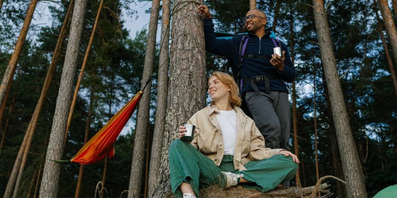 Couple camping A young Black man with a bald head wearing  and a young white woman with red hair wear camping clothes and drink coffee from metal mugs at their woodland campsite, smiling as they look at something off camera.