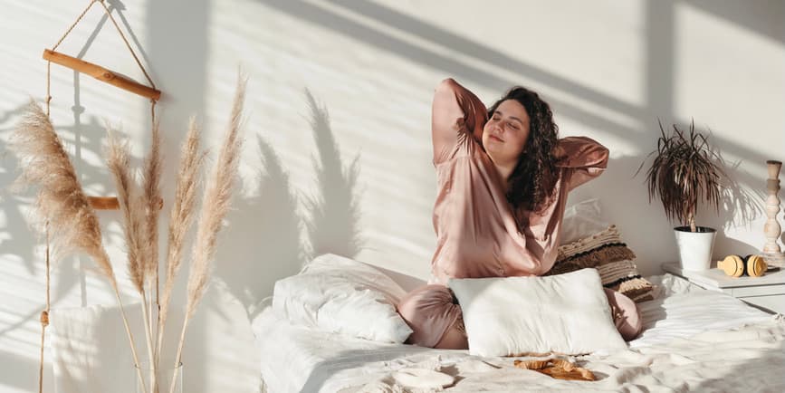 Una joven adulta que se despierta con un pijama de seda pastel se estira después de dormir y disfruta de una mañana soleada en un acogedor ambiente hogareño.
