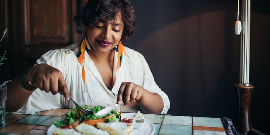 Una hermosa mujer india vestida con una camisa blanca y pendientes largos de plumas se sienta en la mesa de un restaurante y sonríe mientras corta un plato colorido.