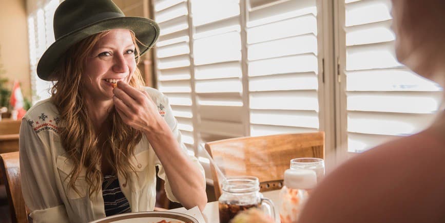 Woman eating at diner A young white woman with long wavy blond hair wearing a fedora and button down shirt smiles as she eats a french fry while sitting at a diner booth next to a window with sun streaming through.