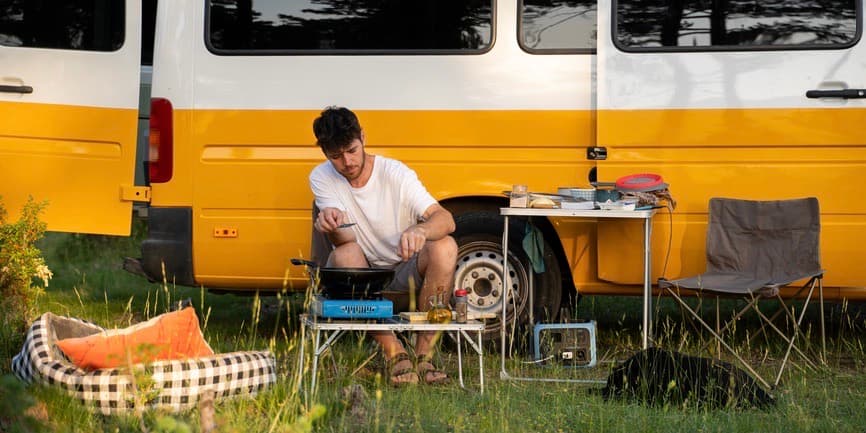 Man camping with van An outdoor summery photo of a young white man sitting in front of a yellow and white camper van, making food at a camping stove with a table of equipment nearby and a dog bed and black dog lying in the grass.