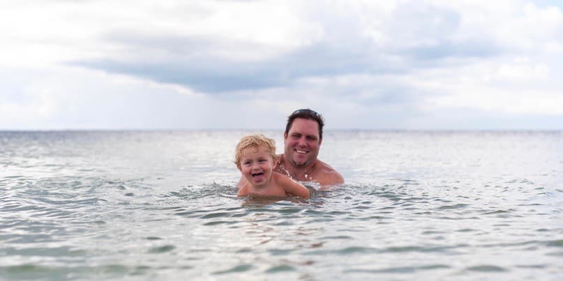 Father and son in ocean. A young white toddler with blond hair learns how to swim in the Caribbean Sea with their father, a smiling white man with brown hair.