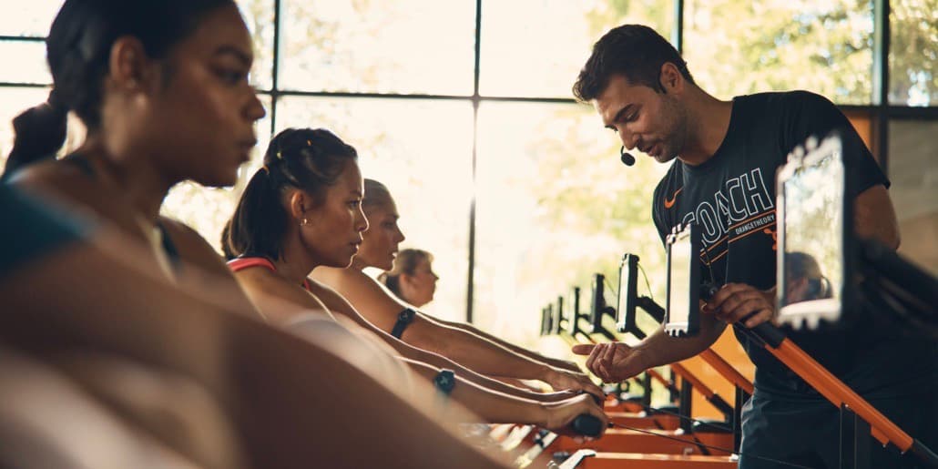 An Orangetheory Coach encourages a young woman on a rowing machine during an Orangetheory Fitness class. An Orangetheory Coach encourages a young woman on a rowing machine during an Orangetheory Fitness class.
