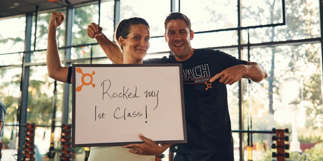 A middle-aged white woman smiles, makes a muscle and and holds a sign that says \"Rocked my 1st class\" while standing next to a white male Orangetheory Coach who also smiles and points down at the sign. A middle-aged white woman smiles, makes a muscle and and holds a sign that says "Rocked my 1st class" while standing next to a white male Orangetheory Coach who also smiles and points down at the sign.