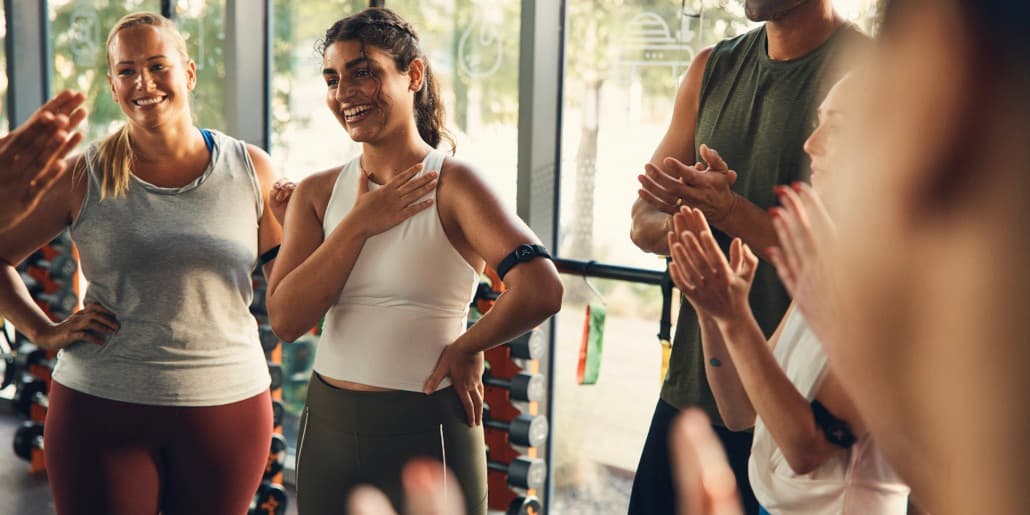 A young brown-skinned woman with brown hair smiles as she's being applauded by Orangetheory Fitness classmates. A young brown-skinned woman with brown hair smiles as she's being applauded by Orangetheory Fitness classmates.