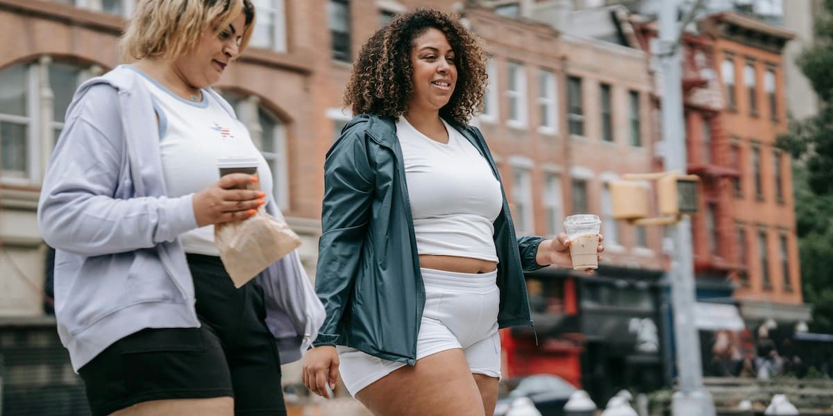 Dos amigas con cuerpos más grandes caminan por la ciudad de Nueva York en ropa deportiva después de correr, cargando cafés mientras sonríen y hablan.