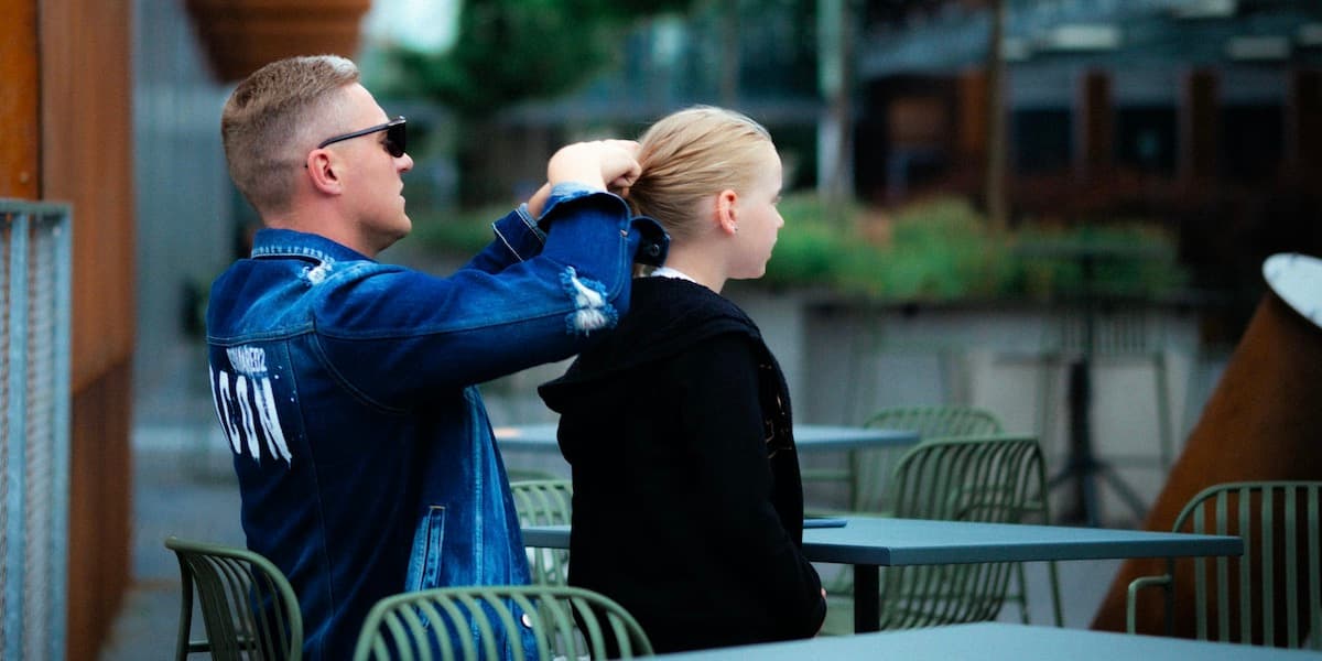 Father doing daughter's hair outside A white father with blond hair wearing a blue jersey sits outside at otherwise empty metal tables, braiding the blond hair of his young daughter.