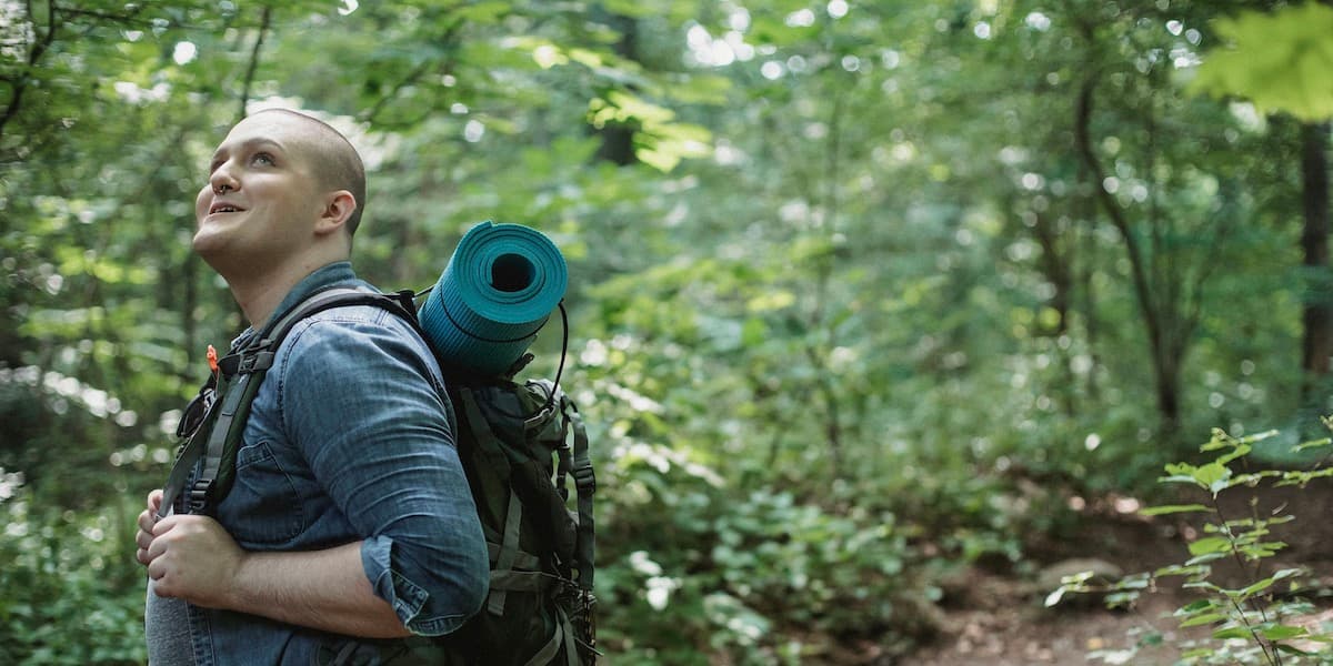 Man hiking and smiling at trees A male hiker with no hair wearing a blue shirt and a backpack with a yoga mat on top smiles as he looks up at something in the trees.