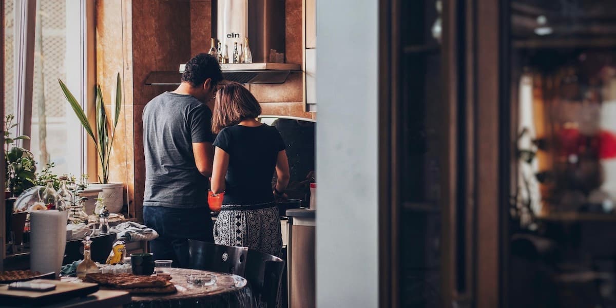 Una pareja blanca permanece junta junto a la estufa en una cocina cálida y desordenada, con muchos detalles en madera y un cómodo desorden.