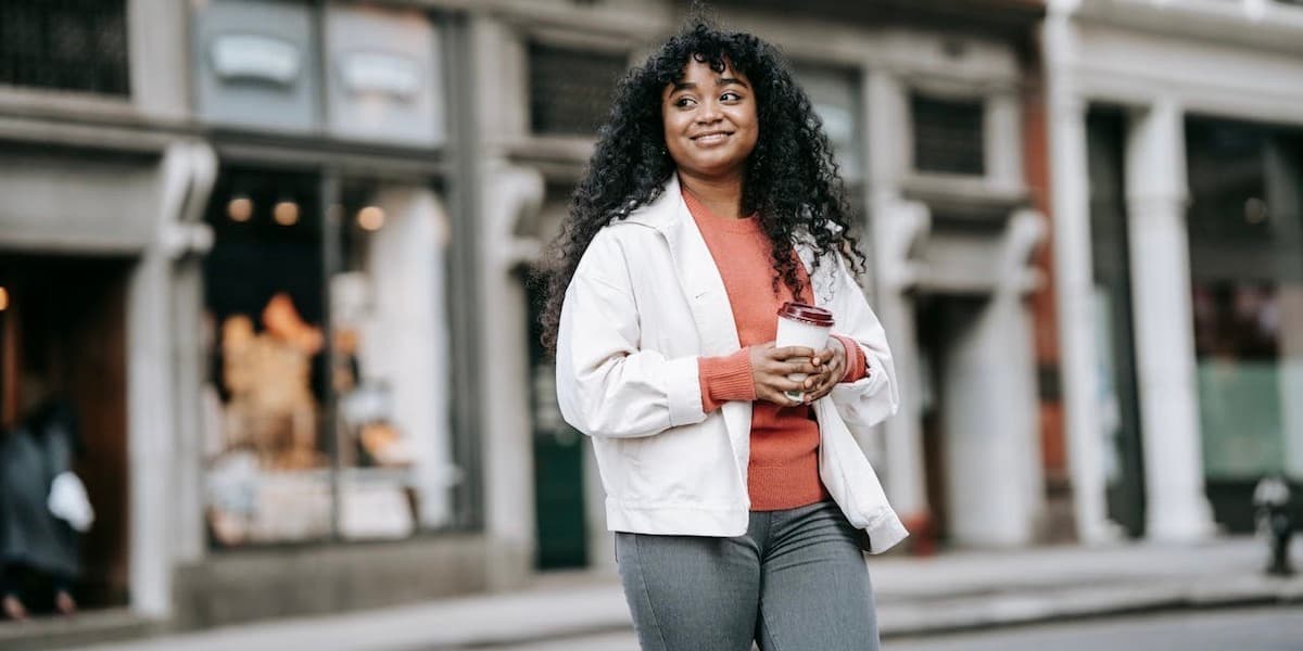 Woman walking on city street Cheerful young Black woman wearing casual clothing with takeaway coffee walking on street.