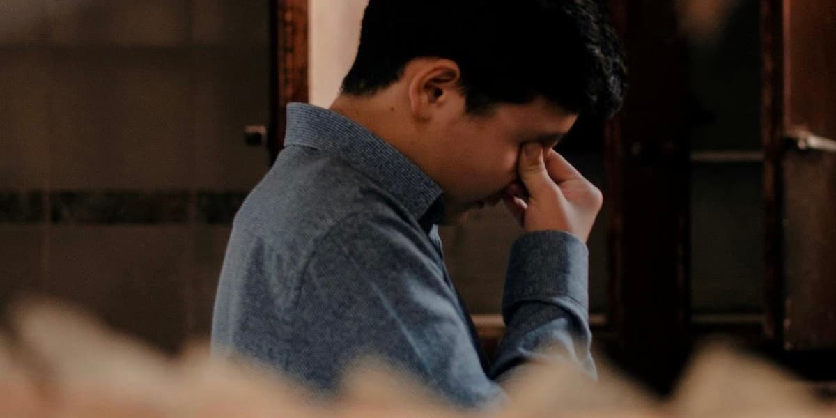 Man with headache A young Hispanic man with black hair wearing a denim shirt is seen through shelves with his head bent down, pinching his fingers over the bridge of his nose.