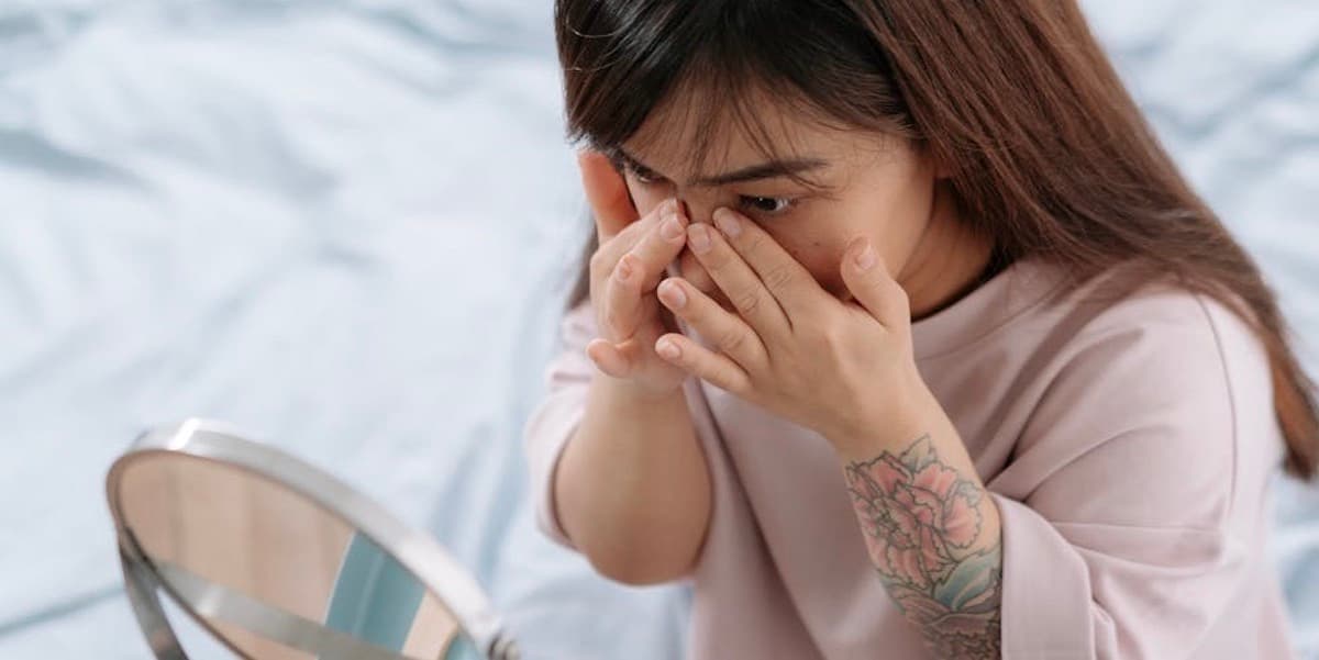 Woman putting on moisturizer A female little person with long brown hair wearing a pink sweater applies lotion around her eyes while she looks into a mirror that sits on the tabletop in front of her while in a nondescript room.