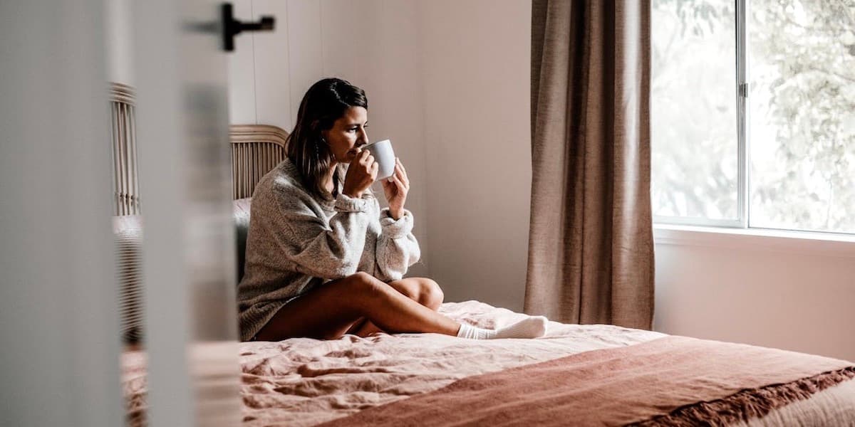 Una joven blanca con cabello largo y castaño oscuro está sentada en su cama con una sudadera y pantalones cortos, bebiendo de una taza.