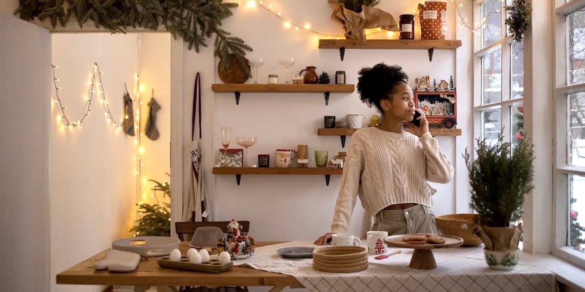 Una joven negra que lleva un jersey habla por teléfono mientras mira por la ventana de su cocina en una acogedora cocina decorada con luces centelleantes, ramas de pino y pequeños árboles. Detrás de ella hay estantes abiertos de pino y la encimera frente a ella está cubierta con material para hornear.