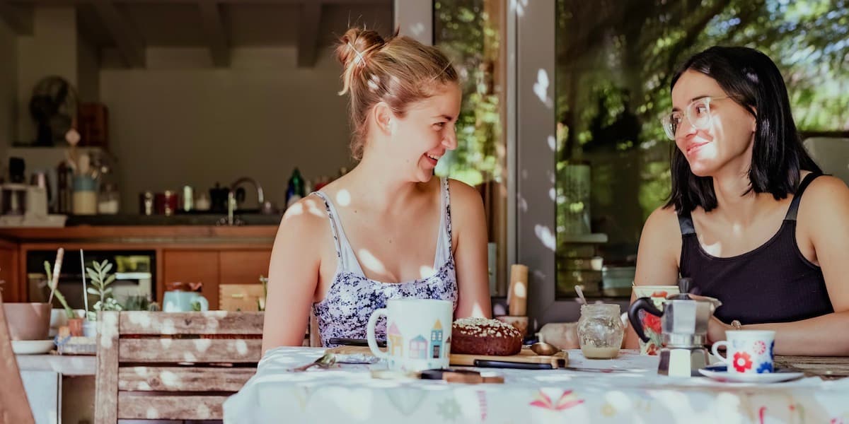 Dos mujeres jóvenes se sientan en una mesa de café al aire libre, bebiendo café exprés y comiendo pastelería, sonriendo mientras conversan.