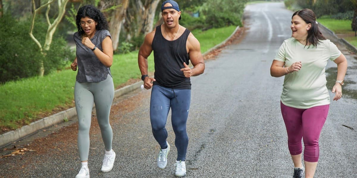 Tres jóvenes de diferentes tamaños y vestidos con ropa deportiva corren juntos por una carretera pavimentada con césped y árboles a un lado.