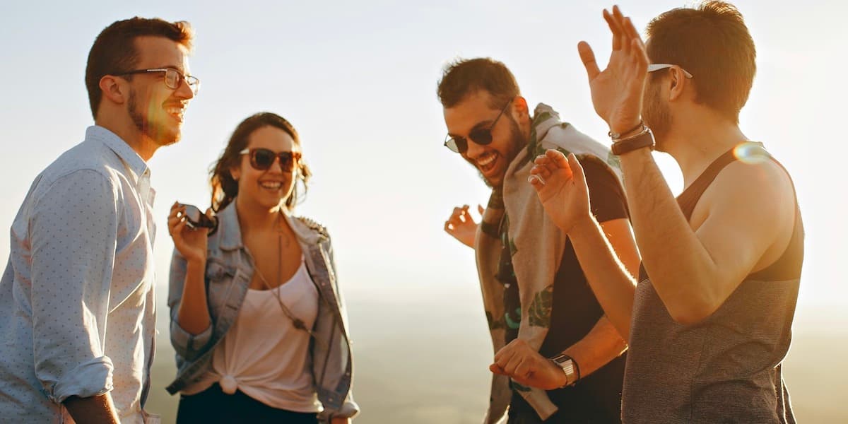 Cuatro jóvenes amigos adultos vestidos con ropa de verano sonríen y ríen en la playa al atardecer, y la luz crea manchas y sombras a su alrededor.