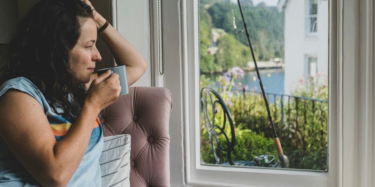 Una mujer, vestida con una colorida camiseta, está sentada en un asiento junto a la ventana de su casa, llevándose una taza a los labios y mirando contemplativamente por la ventana, donde se ve el agua, la vegetación y una caña de pescar apoyada en una silla.