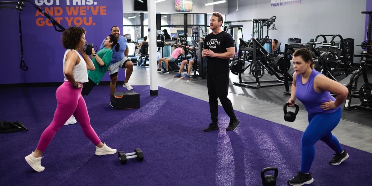 Dos mujeres levantan pesas con la ayuda de un entrenador en Anytime Fitness mientras otras personas hacen ejercicio en segundo plano. Foto de Anytime Fitness.