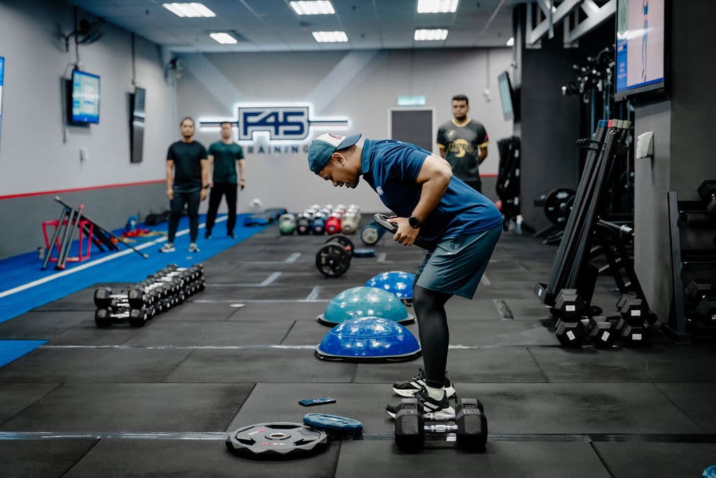 A person performs a bent-over row with a weight plate during an F45 Training session, surrounded by functional training equipment in a gym setting.