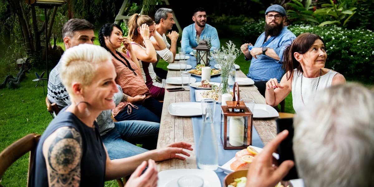 Un grupo de amigos adultos se sientan afuera en una mesa larga de madera cubierta de platos, cubiertos, flores y velas, todos mirando a alguien fuera del marco, para este artículo sobre la microdosificación de GLP-1.