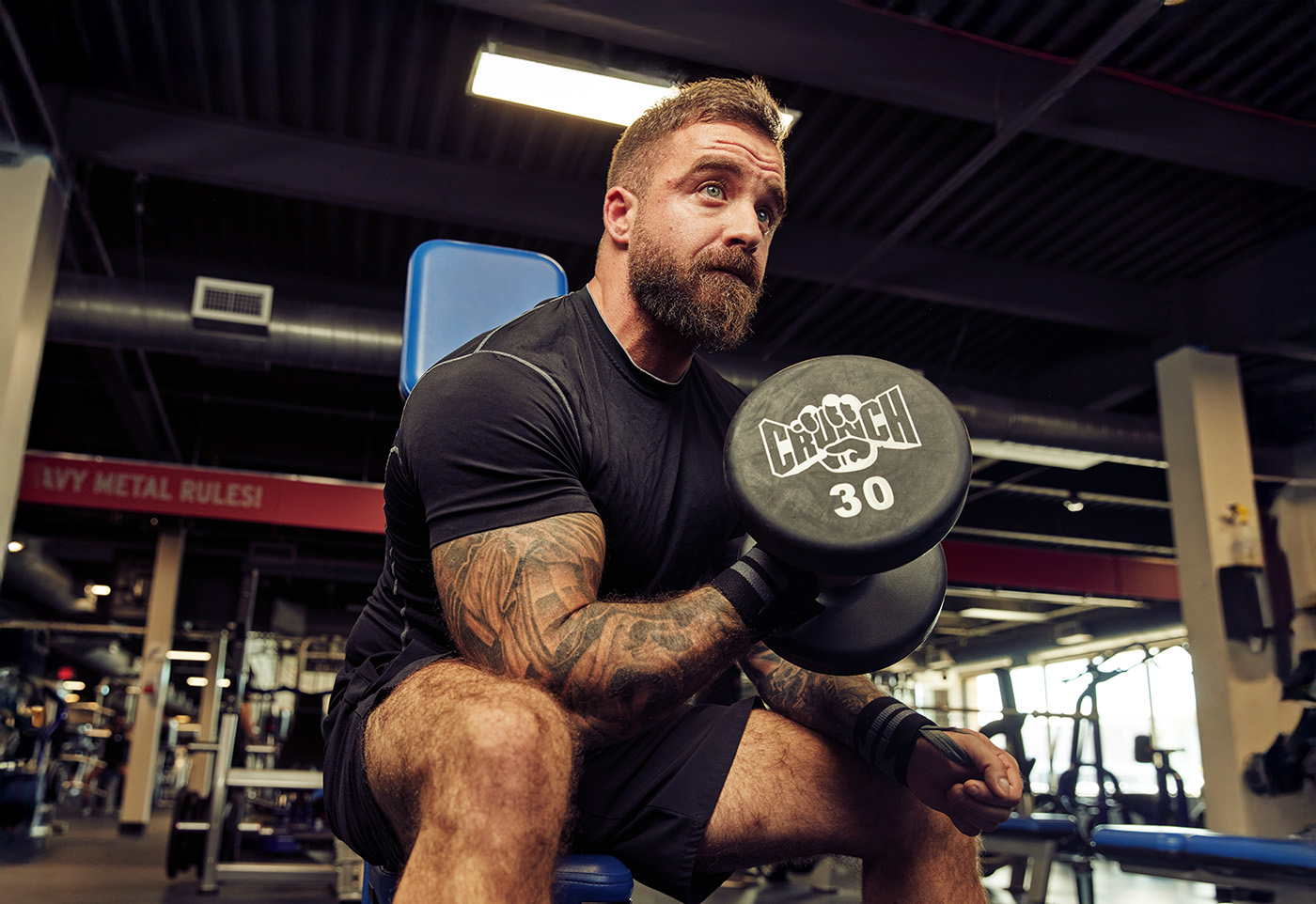 A muscular, bearded man with tattooed arms performs a seated dumbbell curl in a gym, lifting a 30-pound weight while focused on his workout.