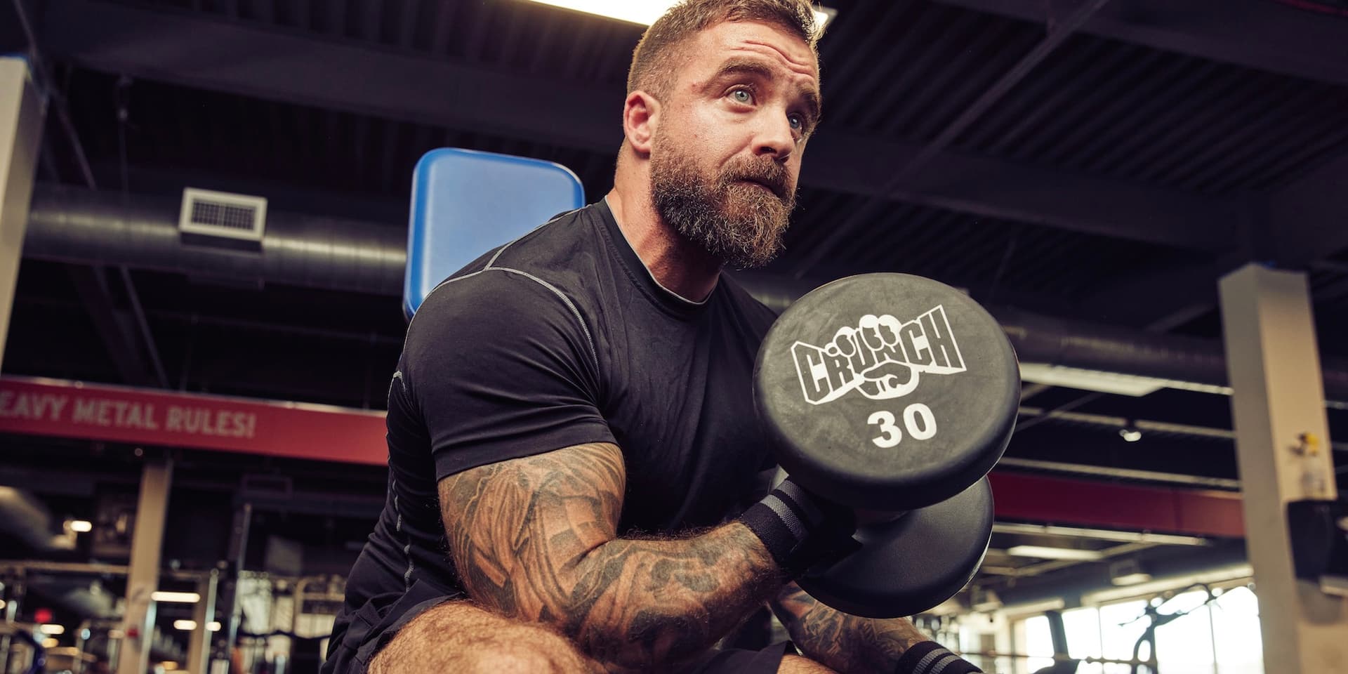Un hombre musculoso y vestido de negro con tatuajes en el brazo se sienta en un banco de trabajo y levanta una mancuerna de 30 libras con el logotipo de Crunch en el extremo en este artículo Dr. B sobre cómo usar su HSA en Crunch.