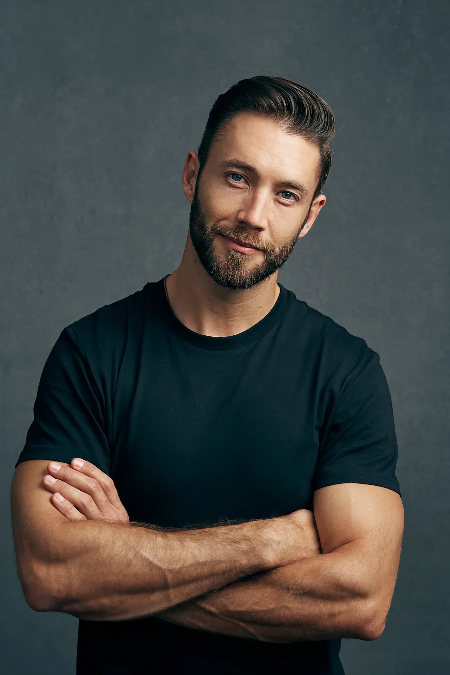 Hombre sonriente con los brazos cruzados en camisa negra de pie frente a un fondo gris
